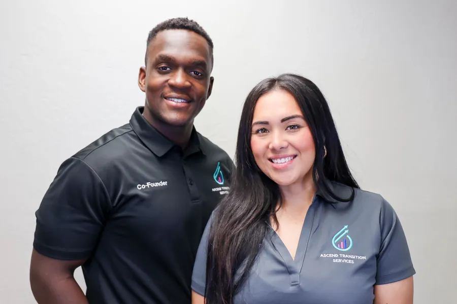 Bridget and Rick Noel, founders of Ascend Transition Services, in matching polo shirts smiling in front of a white wall