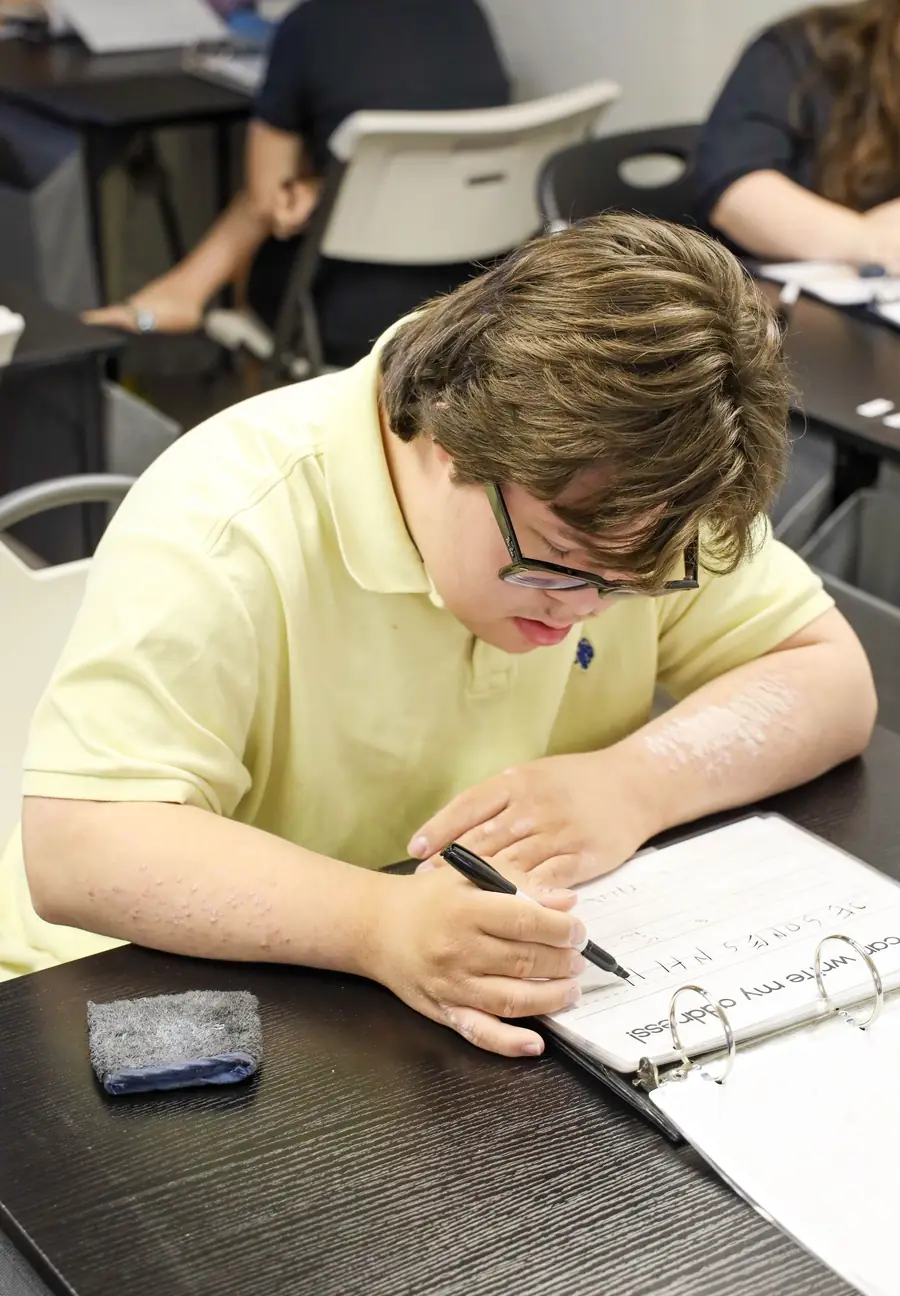 A participant with glasses writing in a notebook at a desk in a classroom setting