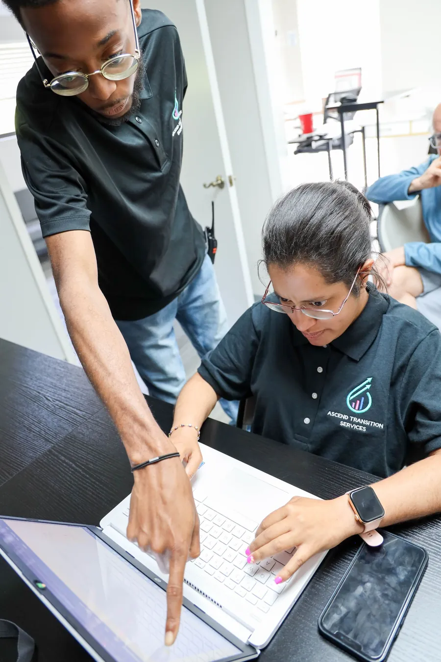 A staff member pointing at a laptop screen while assisting a participant typing at a desk