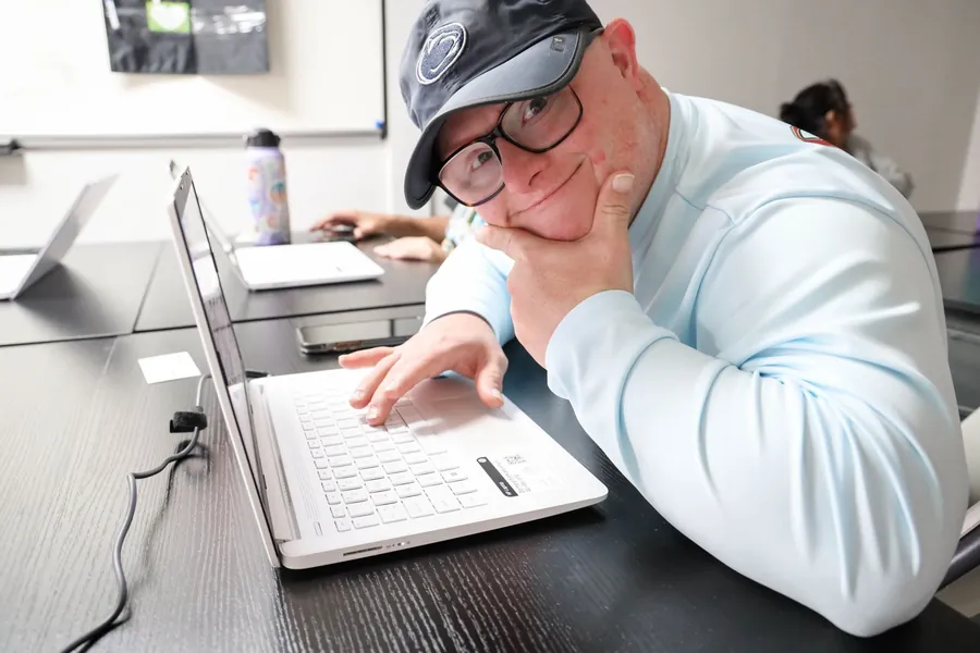 A man wearing glasses and a hat smiling while typing on a laptop at a table indoors